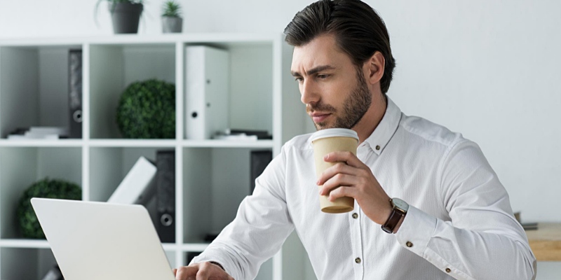 Vista laterale di un ragazzo in camicia bianca che tiene un bicchiere di carta mentre osserva il monitor del Notebook